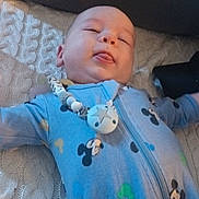 Logan is registered to the contest to win money with this photo: baby, child, infant, blue_onesie, mickey_mouse, pacifier_clip, blanket, knitted, couch, tongue_out, smiling, person, indoor, cute, sleepy, relaxed, face, head, baby_clothing, lying_down
