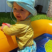Sawyer is registered to the contest to win money with this photo: toddler, child, smiling, hat, swim_trunks, inflatable_pool, water, yellow, blue, outdoor, grass, fence, sunlight, summer, play, happy, person, vacation, fun, backyard