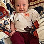 apparel, baby, blanket, child, cozy, cute, face, feet, floral_shirt, hands, happy, indoor, infant, maroon_pants, patterned_blanket, portrait, sitting, smiling, soft_texture, young_child