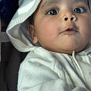 Gowshika is registered to the contest to win money with this photo: baby, child, close_up, curious_expression, cute, eyes, face, hood, indoors, infant, knitwear, mouth, person, portrait, skin, soft_texture, sweater, warm_light, white_clothing, young_child