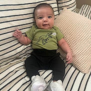 Jael joined the competition — help win amazing prizes! baby, infant, smile, green_shirt, dinosaur_print, white_sneakers, striped_couch, pillows, indoor, cute, happy, child, sitting, casual_clothing, young_baby, portrait, comfortable, home_interior, soft_lighting, adorable