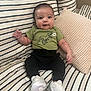 baby, infant, smile, green_shirt, dinosaur_print, white_sneakers, striped_couch, pillows, indoor, cute, happy, child, sitting, casual_clothing, young_baby, portrait, comfortable, home_interior, soft_lighting, adorable