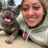 Latto is registered to the contest to win money with this photo: woman, dog, french_bulldog, smiling, happy, outdoor, selfie, tongue_out, curly_hair, piercings, hoodie, chain_collar, concrete, trees, cloudy_sky, face, closeup, pet, friendship, casual