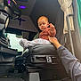 baby, child, red_hair, truck_interior, steering_wheel, seat, hand, curtain, window, vehicle, clothing, person, floor_mat, dashboard, adult_hand, indoors, car_seat, blanket, curtains, blue_eyes