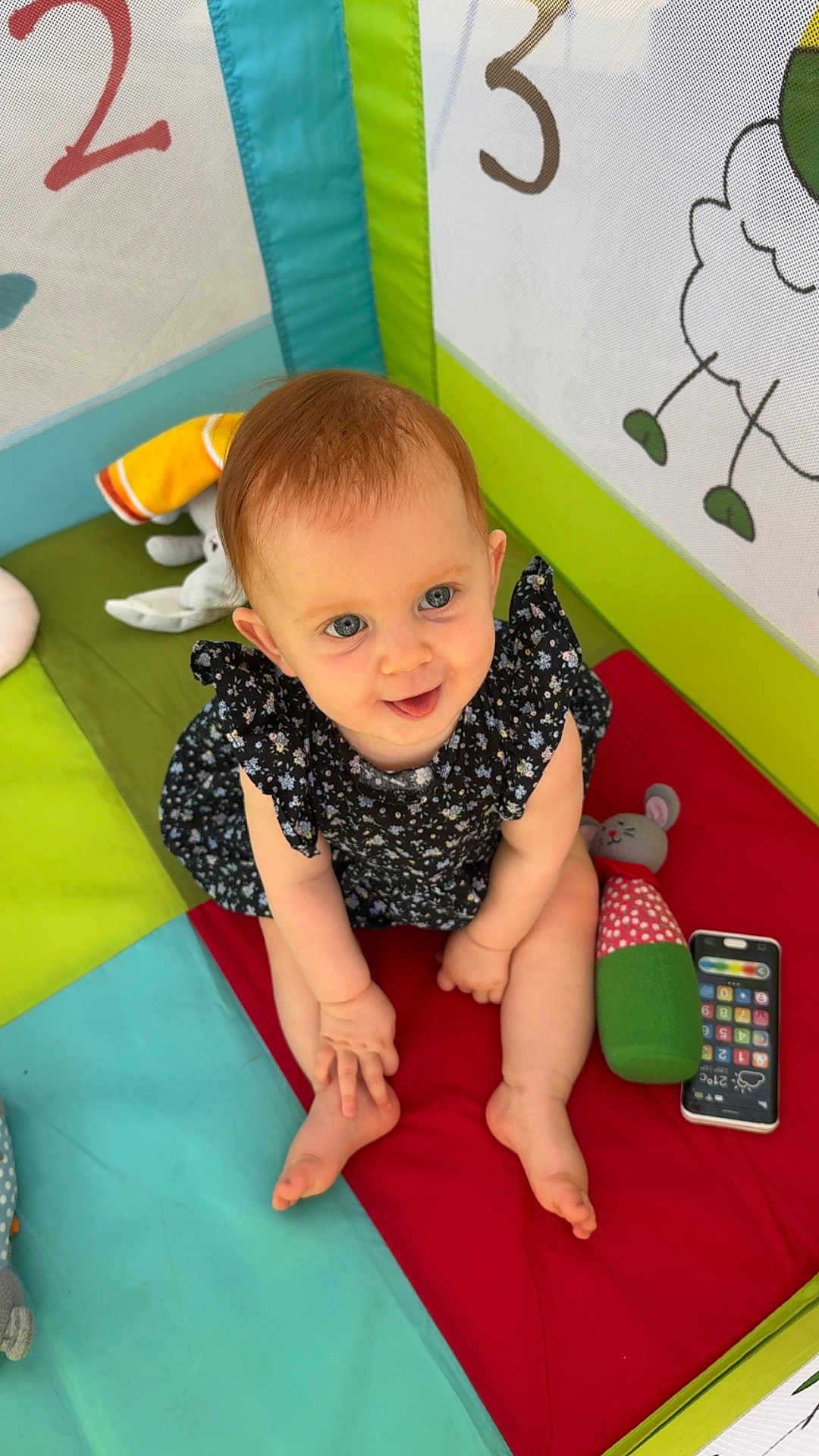 Jöy a rejoint le concours — aidez-le/la à gagner de superbes lots ! baby, child, blue_eyes, sitting, colorful_mat, toy_phone, plush_toy, indoor, playpen, cute, smiling, short_hair, dress, patterned_clothing, soft_toys, numbers, illustration, playful, infant, looking_up