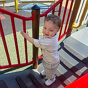 Ronnie is registered to the contest to win money with this photo: casual_clothing, child, climbing, colorful, curly_hair, daylight, fun, outdoor, play_equipment, playground, red_rail, safety_surface, shadow, shorts, smiling, stairs, toddler, tshirt, white_shoes, young_child