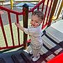 casual_clothing, child, climbing, colorful, curly_hair, daylight, fun, outdoor, play_equipment, playground, red_rail, safety_surface, shadow, shorts, smiling, stairs, toddler, tshirt, white_shoes, young_child
