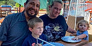 Ryder is registered to the contest to win money with this photo: family, outdoor, table, umbrella, smiling, man, woman, child, boy, toddler, plate, wood, patio, sunlight, casual, happy, green_hair, curly_hair, short_hair, restaurant