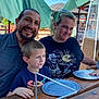 family, outdoor, table, umbrella, smiling, man, woman, child, boy, toddler, plate, wood, patio, sunlight, casual, happy, green_hair, curly_hair, short_hair, restaurant