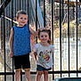 child, children, boy, girl, water_fountain, summer_clothing, smile, outdoor, playful, sunlight, fence, shorts, tshirt, happy, portrait, nature, daytime, casual, park, friends