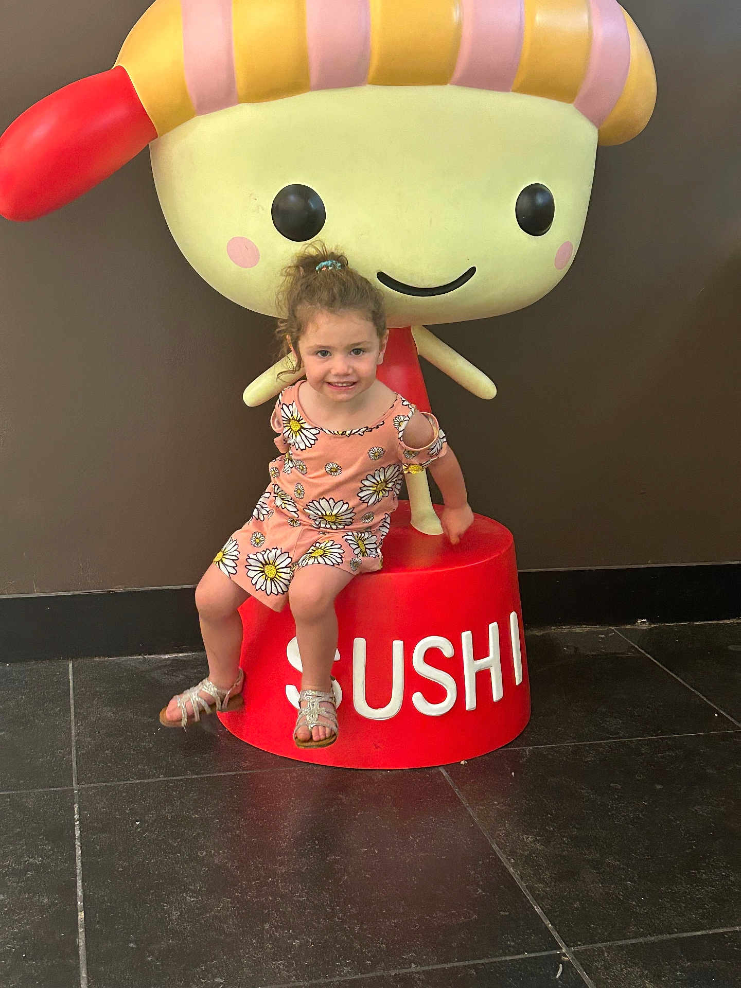 Aurora is registered to the contest to win money with this photo: child, girl, smile, daisy_dress, sandals, sushi, cartoon_character, statue, red_pedestal, floor_tiles, indoor, brown_wall, happy, posing, toy, cute, playful, young, portrait, person