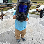Yamari is registered to the contest to win money with this photo: child, welding_helmet, outdoor, concrete, truck, trailer, blue_shirt, tan_pants, sneakers, equipment, helmet, person, standing, yard, grass, vehicle, tool, playful, funny, protective_gear