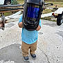 child, welding_helmet, outdoor, concrete, truck, trailer, blue_shirt, tan_pants, sneakers, equipment, helmet, person, standing, yard, grass, vehicle, tool, playful, funny, protective_gear