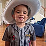 toddler, child, hat, costume, indoor, tongue_out, smiling, spiderman, jersey, person, furniture, living_room, blue_chair, wooden_floor, playful, cute, apparel, young_child, portrait, home