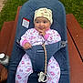 Fernanda is registered to the contest to win money with this photo: baby, infant, smile, onesie, hat, bouncer, babybjorn, pacifier, wooden_table, grass, outdoor, cute, playful, water_bottle, child, seated, happy, person, clothing, face