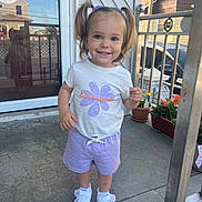 Vianna joined the competition — help win amazing prizes! toddler, child, girl, pigtails, white_tshirt, purple_shorts, white_sneakers, porch, glass_door, reflection, houses, potted_plants, flower, smiling, standing, outdoor, daylight, concrete_floor, railing, happy