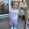 toddler, child, girl, pigtails, white_tshirt, purple_shorts, white_sneakers, porch, glass_door, reflection, houses, potted_plants, flower, smiling, standing, outdoor, daylight, concrete_floor, railing, happy
