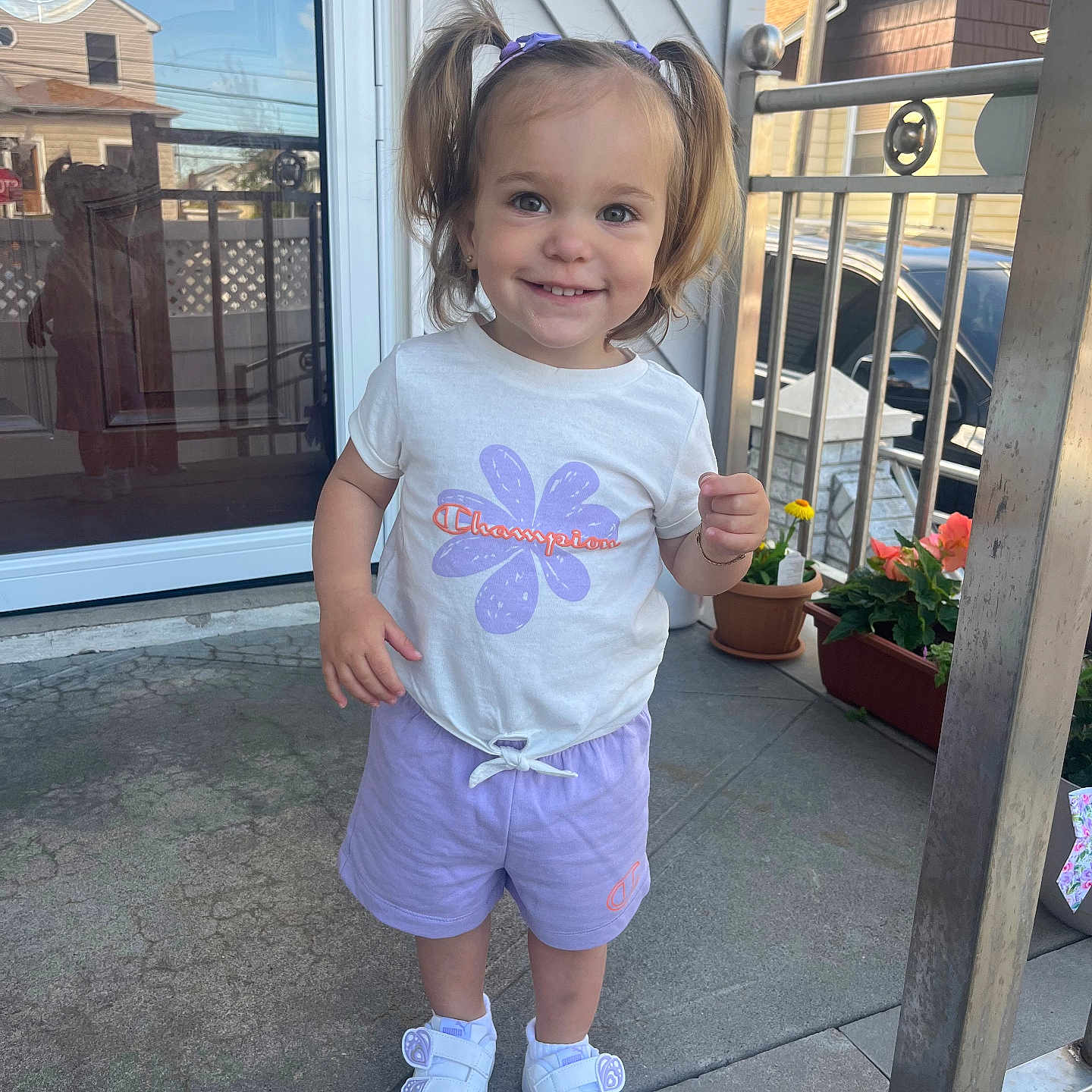 Vianna joined the competition — help win amazing prizes! toddler, child, girl, pigtails, white_tshirt, purple_shorts, white_sneakers, porch, glass_door, reflection, houses, potted_plants, flower, smiling, standing, outdoor, daylight, concrete_floor, railing, happy