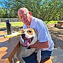 man, dog, outdoor, picnic_table, glasses, white_shirt, jeans, sunlight, trees, grass, happy_dog, embrace, pet, nature, daylight, bench, smile, leash, fence, shade