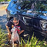 man, dog, vehicle, car, outdoor, sunlight, plants, parking_lot, black_suv, sports_logo, raiders, glasses, tattoos, pet, crouching, leash, greenery, shadow, casual_clothing, daytime
