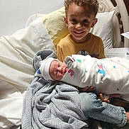 Maverick joined the competition — help win amazing prizes! child, baby, hospital_bed, blanket, swaddled, smiling, curly_hair, pillow, clothing, newborn, person, indoors, footwear, jeans, happy, family, portrait, hospital_room, bed, toddler