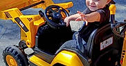 Marcelino is registered to the contest to win money with this photo: child, toddler, boy, smiling, outdoor, toy_vehicle, toy_loader, construction, excavator, yellow, black, steering_wheel, driving, seated, baseball_cap, casual_clothing, pavement, sunlight, portrait, playtime