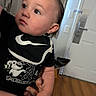 baby, infant, child, portrait, face, looking_up, curious, indoors, bathroom, door, floor, wood_floor, adult_hand, black_clothing, bib, cartoon, skeleton, graphic_shirt, hair, candid