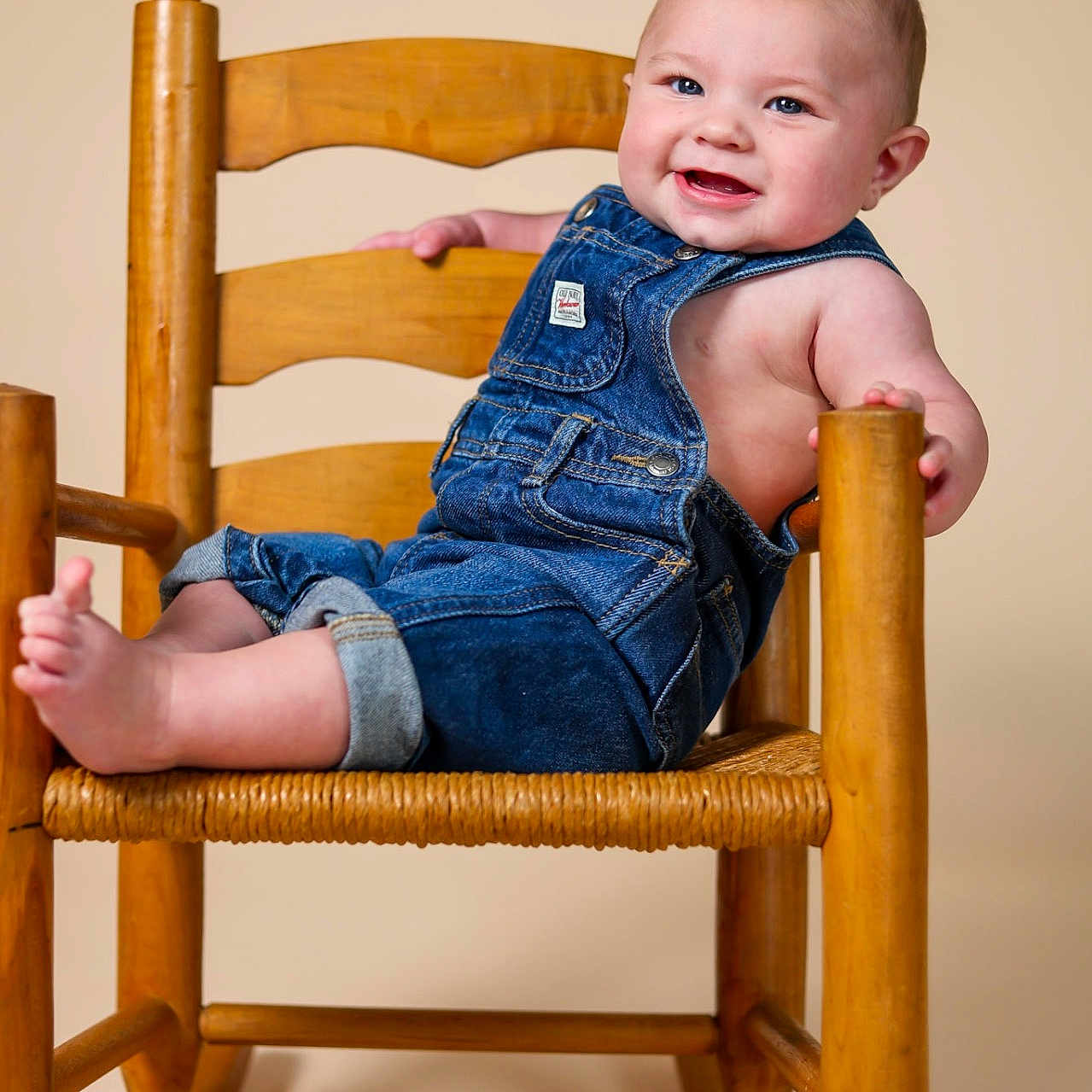 Riley is registered to the contest to win money with this photo: baby, child, smile, denim_overalls, rocking_chair, wooden_chair, happy, cute, infant, portrait, sitting, foot, hand, blue_eyes, studio, indoors, clothing, person, furniture, toddler