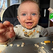 Jensen is registered to the contest to win money with this photo: baby, toddler, child, highchair, messy, food_smear, blue_eyes, smile, teeth, reaching_hand, tray, snack, cereal, puree_pouch, bib, indoor, window, portrait, closeup, happy
