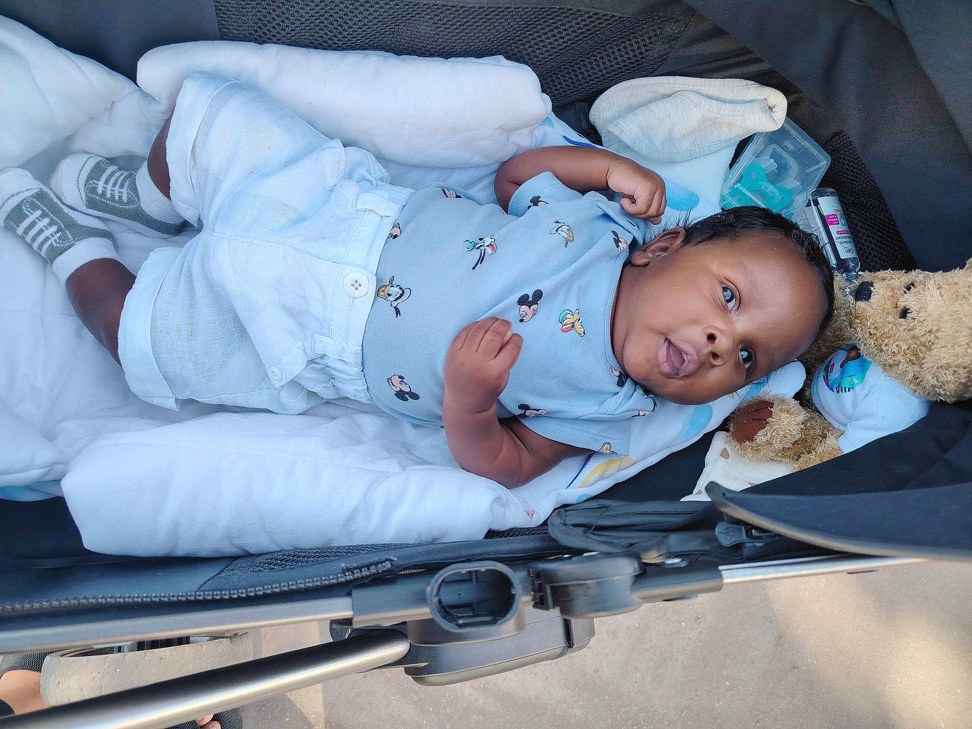 Djawël participe au concours pour gagner de l'argent avec cette photo : baby, infant, stroller, blanket, teddy_bear, clothing, mickey_mouse, socks, cute, child, lying_down, comfort, soft_toys, outdoor, sunlight, person, young, portrait, resting, cozy