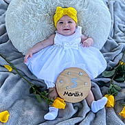Imogen is registered to the contest to win money with this photo: baby, infant, child, white_dress, yellow_headband, smiling, pillow, blanket, outdoor, grass, yellow_roses, flowers, wooden_sign, celebration, milestone, happy, cute, socks, person, portrait