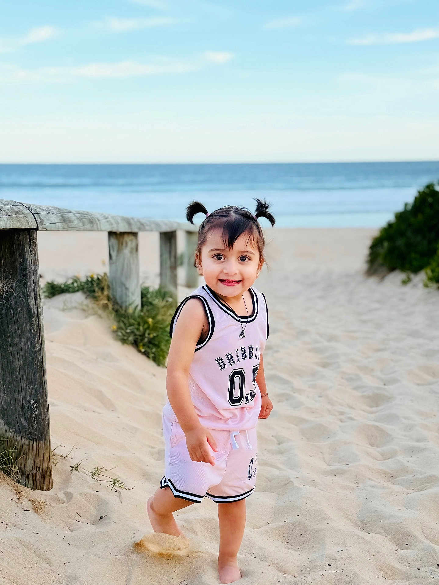 Yamini is registered to the contest to win money with this photo: barefoot, beach, child, daylight, happy, nature, ocean, outdoor, pigtails, pink_outfit, playful, portrait, sand, sky, smile, sporty_clothing, summer, toddler, wooden_fence, young_child