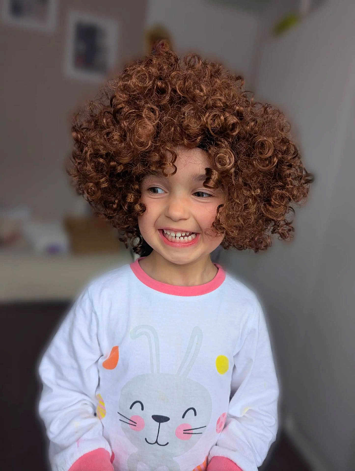 Sophia joined the competition — help win amazing prizes! child, curly_hair, smiling, pajamas, indoor, cute, happy, face, person, childhood, playful, curly_wig, long_sleeve, white_clothing, pink_trim, bunny_design, cheerful, portrait, young, fun