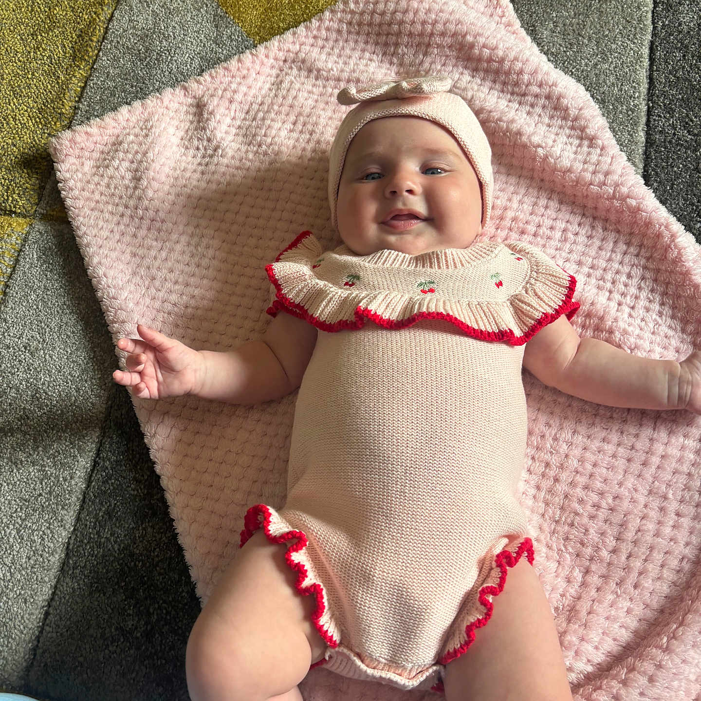 Truley is registered to the contest to win money with this photo: baby, carpet, cheeks, child, cute, floor, happy, headwear, indoors, infant, knitted_outfit, laying_down, legs, patterned_floor, pink_blanket, portrait, red_trim, small_hands, smiling, soft_texture