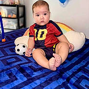 Matias joined the competition — help win amazing prizes! baby, child, soccer_ball, blue_blanket, rainbow_pillow, blue_balloon, sporty_clothing, number_10, indoor, curious_expression, cute, sitting, soft_lighting, home, toy, infant, portrait, playful, feet, face