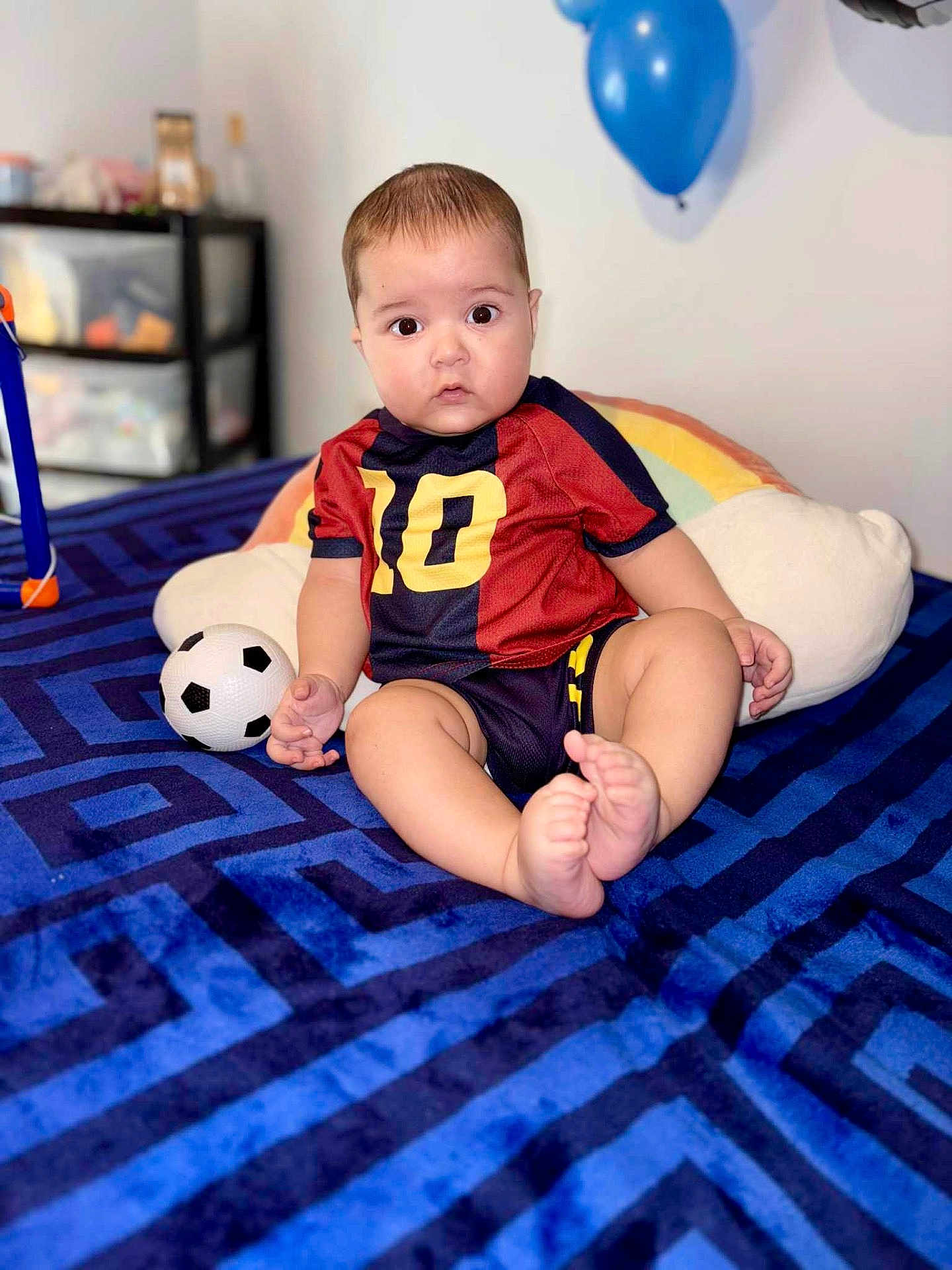 Matias joined the competition — help win amazing prizes! baby, child, soccer_ball, blue_blanket, rainbow_pillow, blue_balloon, sporty_clothing, number_10, indoor, curious_expression, cute, sitting, soft_lighting, home, toy, infant, portrait, playful, feet, face