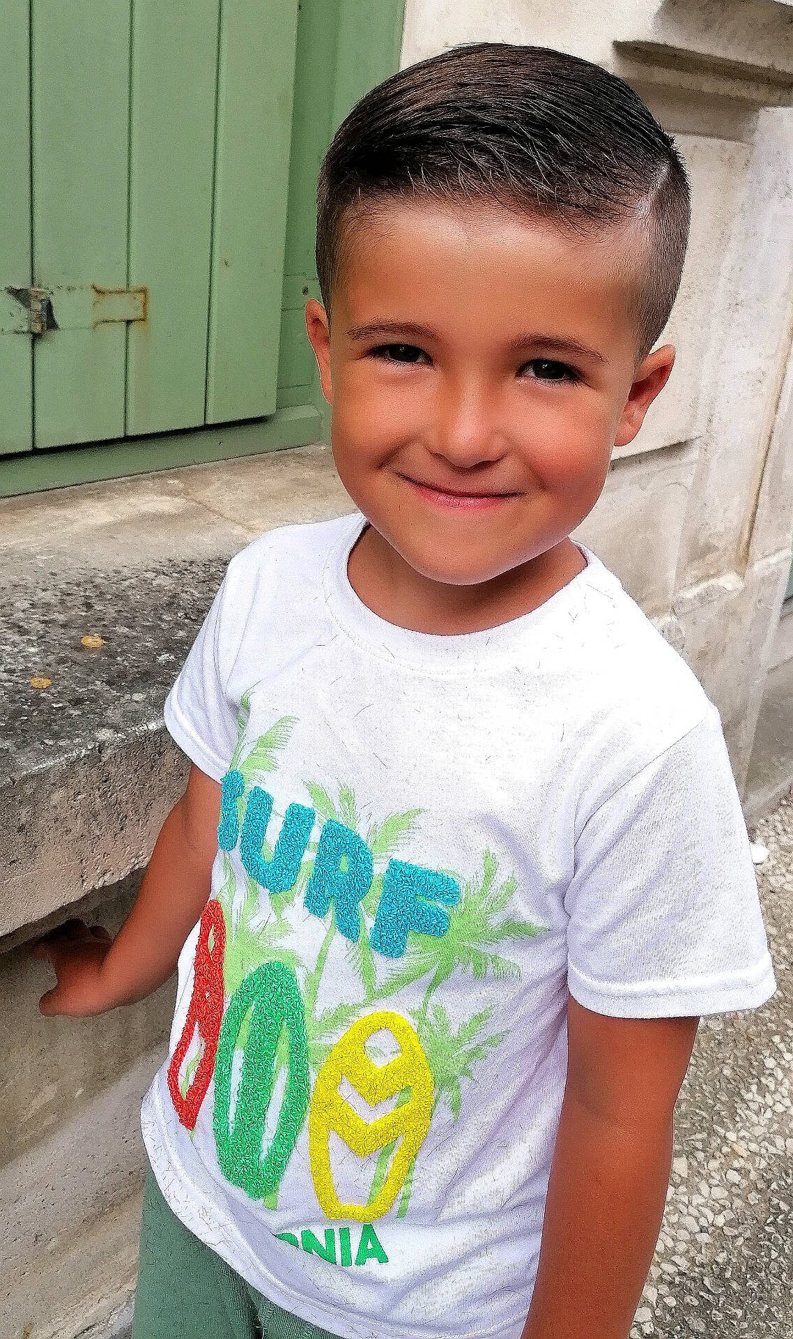 Nathan a rejoint le concours — aidez-le/la à gagner de superbes lots ! background, boy, casual, child, colorful_clothing, concrete, daylight, green_shutter, happy, outdoor, person, portrait, short_hair, smile, standing, stone_wall, tshirt_print, urban, white_tshirt, young