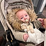 baby, stroller, blanket, pacifier, teddy_bear, fur_hood, hand, jacket, indoor, carpet, person, child, comfort, warm_clothing, infant, toy, holding_hand, cute, portrait, seated