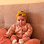 baby, infant, child, headband, bow, couch, pillow, blanket, onesie, socks, sitting, portrait, indoor, soft_texture, pink_clothing, mustard_yellow, big_eyes, chubby_cheeks, buttoned_cardigan, cozy