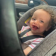 Ryder is registered to the contest to win money with this photo: toddler, child, car_seat, smiling, curly_hair, seatbelt, car_interior, window, warning_label, happy, baby, person, sitting, clothing, face, head, transportation, daylight, cute, infant