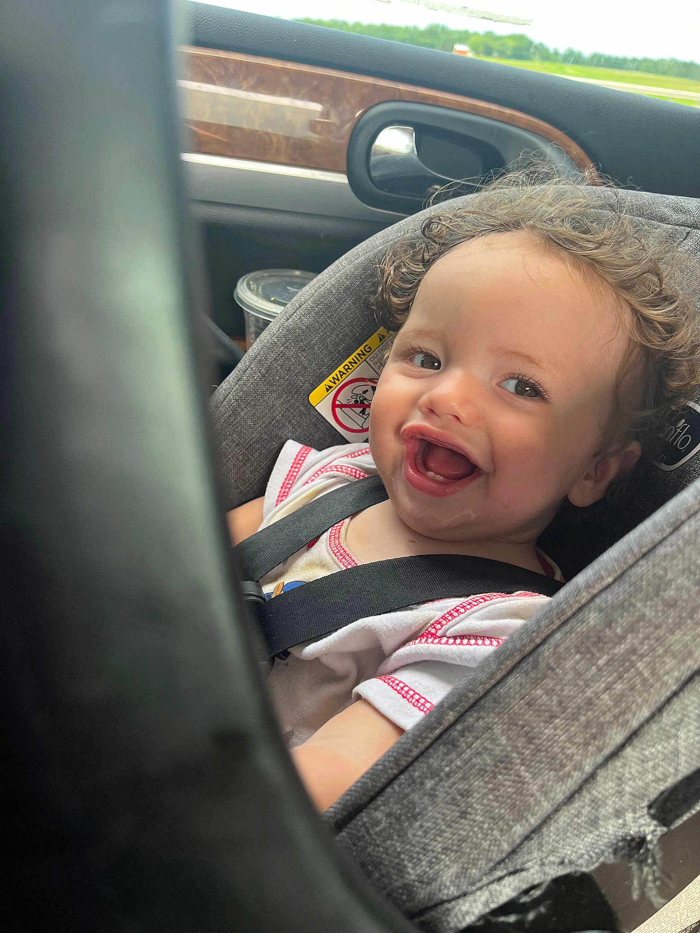Ryder is registered to the contest to win money with this photo: toddler, child, car_seat, smiling, curly_hair, seatbelt, car_interior, window, warning_label, happy, baby, person, sitting, clothing, face, head, transportation, daylight, cute, infant