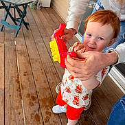 Cain is registered to the contest to win money with this photo: toddler, child, toy, red_hair, shirt, shorts, sneakers, adult_hands, wooden_deck, folding_chair, folding_table, outdoor, window, door, person, playtime, smile, curious, casual_clothing, daylight