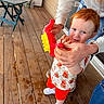 toddler, child, toy, red_hair, shirt, shorts, sneakers, adult_hands, wooden_deck, folding_chair, folding_table, outdoor, window, door, person, playtime, smile, curious, casual_clothing, daylight