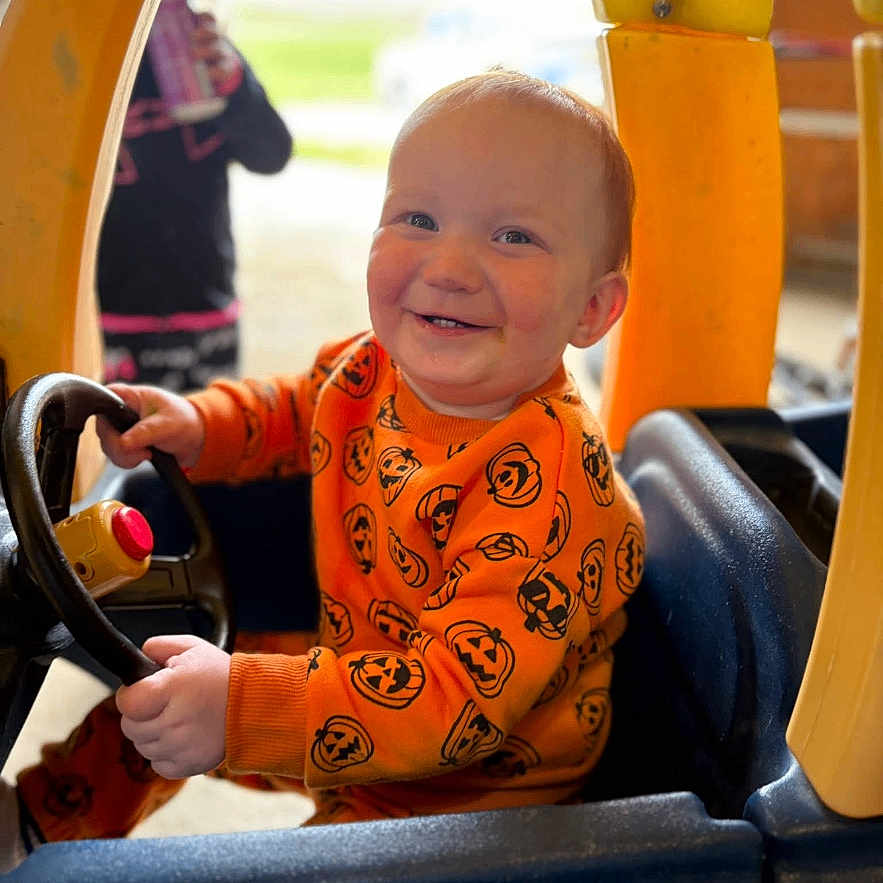 Cain is registered to the contest to win money with this photo: toddler, child, smiling, orange_sweater, toy_car, steering_wheel, indoor, playtime, happy, person, plastic, seat, window, hand, face, cute, young_child, casual_clothing, bright_colors, fun
