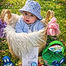 baby, basket, blue_clothing, child, chocolate, cute, easter_toys, face, flowers, grass, greenery, hat, nature, outdoor, person, playful, plush, smiling, spring, sunlight
