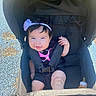 baby, stroller, child, smiling, headband, flower, baby_bottle, black_clothing, outdoor, sunlight, happy, infant, seatbelt, baby_girl, person, cute, portrait, baby_face, sitting, playground_surface