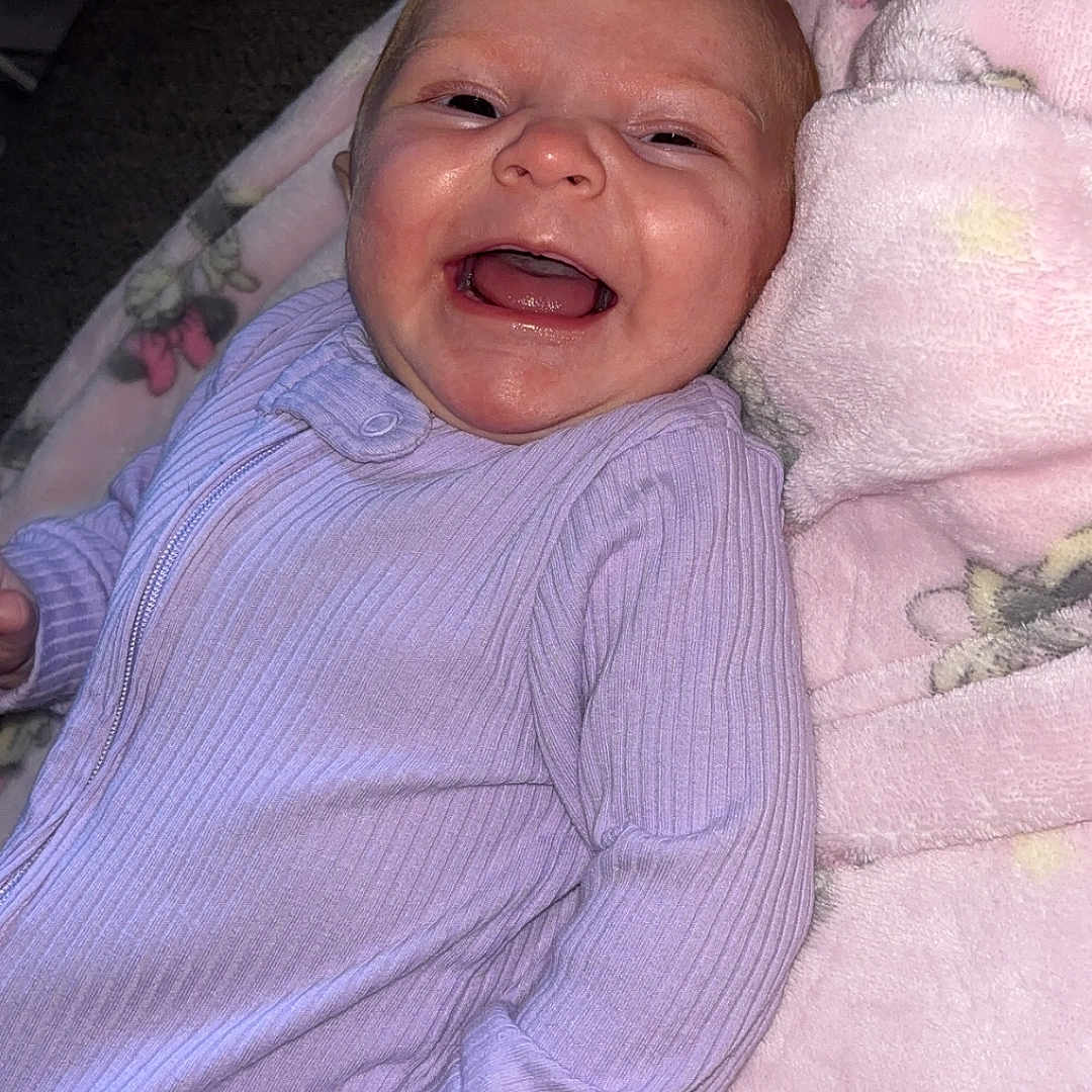Myla joined the competition — help win amazing prizes! baby, infant, smiling, happy, onesie, purple_clothing, blanket, pink_blanket, floral_pattern, cozy, cute, lying_down, face, skin, child, newborn, portrait, indoor, soft_texture, comfort