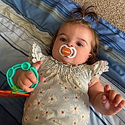 Oakley is registered to the contest to win money with this photo: baby, infant, child, portrait, lying_down, pacifier, toy, floral_outfit, dress, striped_bedding, bed, indoors, brown_hair, ponytail, face, eyes, hand, smiling, close_up, safe