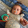 baby, infant, child, portrait, lying_down, pacifier, toy, floral_outfit, dress, striped_bedding, bed, indoors, brown_hair, ponytail, face, eyes, hand, smiling, close_up, safe