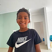 Salim participe au concours pour gagner de l'argent avec cette photo : black_tshirt, boy, bright_lighting, casual_clothing, chair, child, curly_hair, doorway, floor, green_wall, happy, indoor, modern_room, nike_logo, person, portrait, smiling, standing, white_wall, young